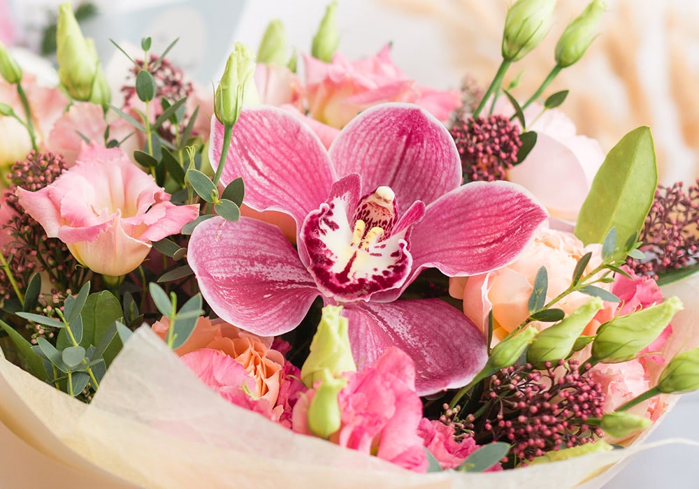 A pink orchid centerpiece sits amidst an arrangement of various flowers, including roses and greenery, all gathered in a bouquet with light-colored wrapping.