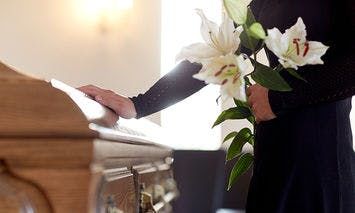 A person dressed in black holds a bouquet of white lilies and rests a hand on a closed wooden coffin in a softly lit room.