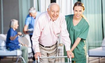 An elderly man uses a walker, assisted by a female caregiver in green scrubs, in an outdoor setting where other elderly individuals are also being attended to.